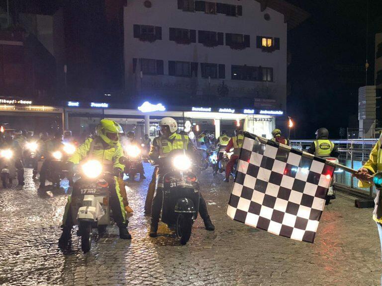Viele Vespas von vorne im Dunklen vor einem Haus - eine Startfahne wird davor geschwenkt