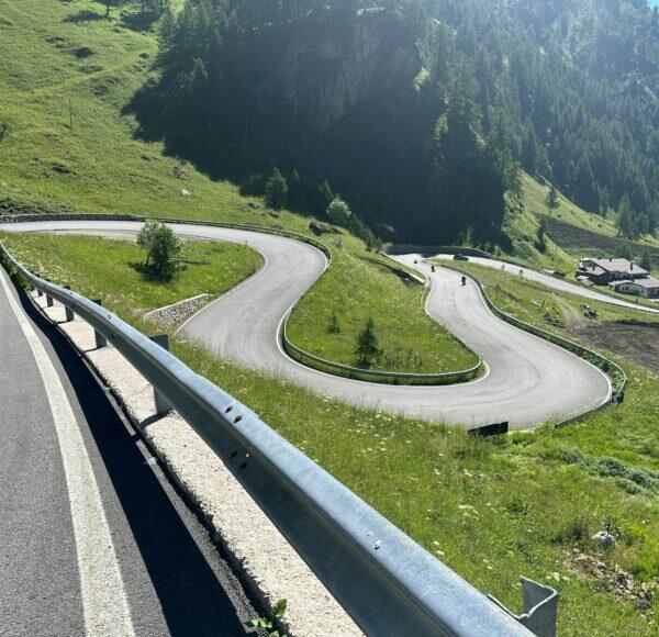 Kurvige Bergstraße mit Leitplanken, umgeben von grünem Gras und Bäumen, sonniges Wetter.