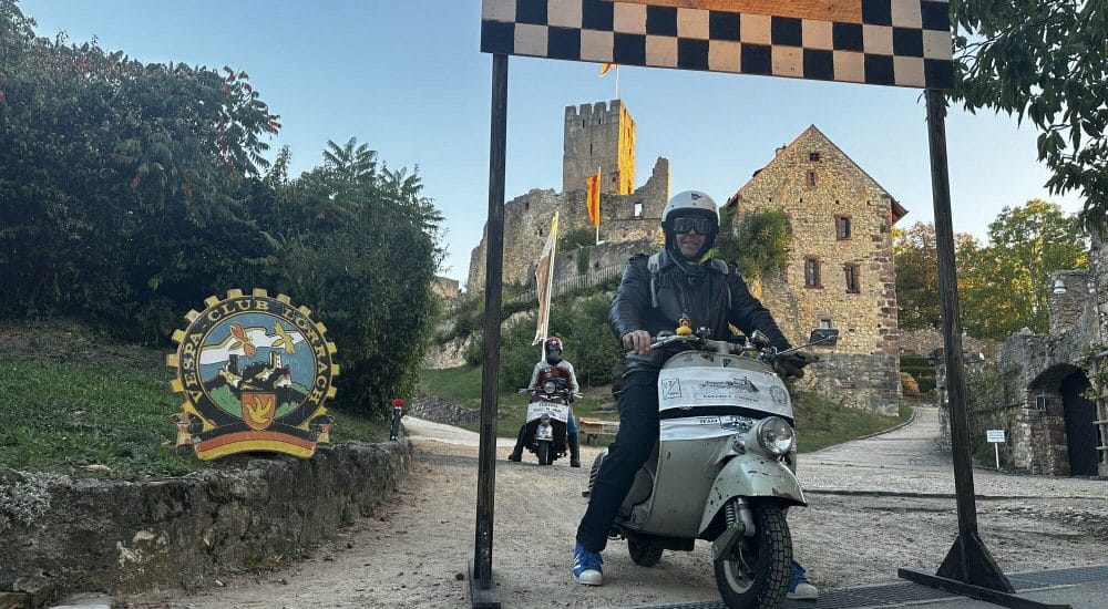 Person fährt Motorroller durch ein Start-Ziel-Tor vor einer Burg, weitere Person folgt, Umgebung ländlich und sonnig.