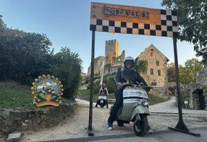 Person fährt Motorroller durch ein Start-Ziel-Tor vor einer Burg, weitere Person folgt, Umgebung ländlich und sonnig.