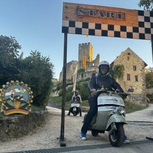 Person fährt Motorroller durch ein Start-Ziel-Tor vor einer Burg, weitere Person folgt, Umgebung ländlich und sonnig.