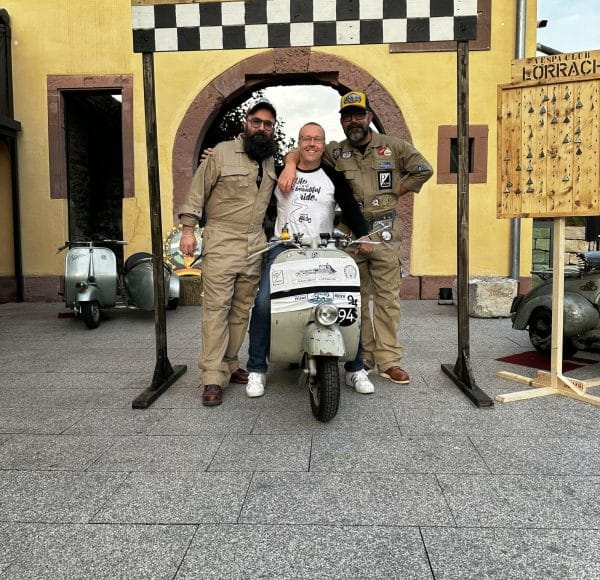 Drei Personen posieren vor einem Startbogen, eine sitzt auf einem Motorroller, die anderen stehen daneben. Im Hintergrund gelbes Gebäude und weitere Roller.