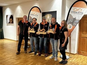 Fünf Personen posieren drinnen, drei halten Holzpokale mit Unendlichkeitssymbol, alle tragen schwarze Jacken mit orangefarbenen Details.