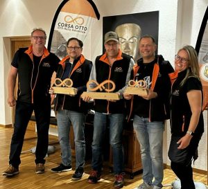 Fünf Personen posieren drinnen, drei halten Holzpokale mit Unendlichkeitssymbol, alle tragen schwarze Jacken mit orangefarbenen Details.