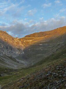 Sonnenbeleuchtete Serpentinenstraße schlängelt sich durch ein steiles Bergtal unter blau bewölktem Himmel.