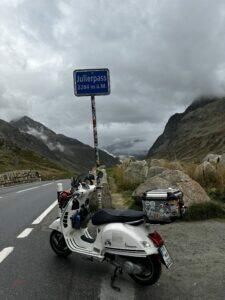 Motorrad an einer kurvigen Bergstraße mit wolkigem Himmel und Passschild, umgeben von Bergen und felsiger Landschaft.