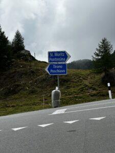 Blaues Straßenschild mit Wegweisern nach St. Moritz, Pontresina, Tirano und Poschiavo, an einer kurvigen Landstraße, Bäume und Hügel im Hintergrund.