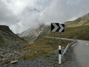 Bergstraße mit Kurvenschild, umgeben von bewölkten Bergen und grüner Landschaft.
