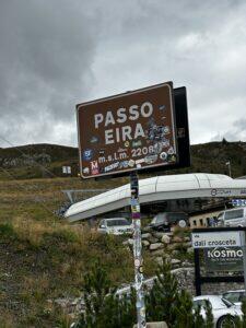 Braunes Schild mit der Aufschrift „Passo Eira, 2208 m ü. M.“ in den Bergen, Autos und Gebäude im Hintergrund, bewölkter Himmel.