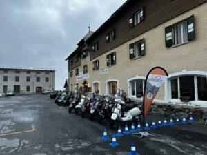 Mehrere Motorroller sind vor einem großen, steinernen Gebäude mit Fensterläden geparkt, daneben steht ein orangefarbenes Werbebanner, Himmel ist bewölkt.