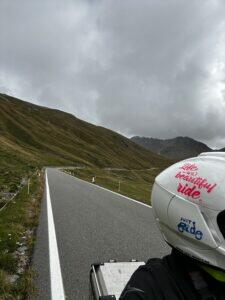 Leere Straße durch bergige Landschaft, Motorradhelm am rechten Bildrand, bewölkter Himmel.