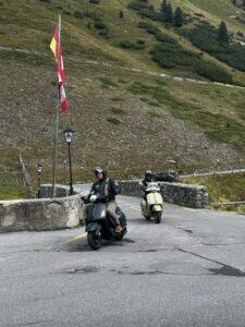 Zwei Personen fahren Motorroller auf einer kurvigen Bergstraße, umgeben von grüner Landschaft und Fahnen im Hintergrund.