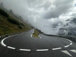 Kurvige Bergstraße mit nassem Asphalt, umgeben von Nebel und bewaldeten Hängen.