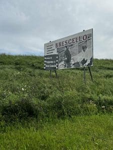 Großes Brescello-Schild mit Schwarzweißfoto, das zwei Personen auf einem Motorrad zeigt, steht auf einer grünen Wiese unter wolkigem Himmel.