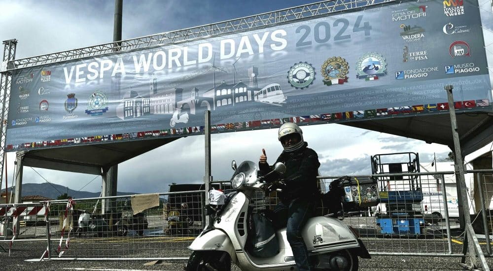 Olaf H. Just am Eingang und vor dem Banner der Vespa World Days 2024 auf einer weißen Vespa GTS 300 sitzend.