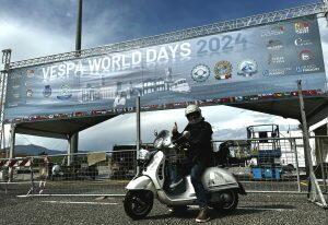 Olaf H. Just am Eingang und vor dem Banner der Vespa World Days 2024 auf einer weißen Vespa GTS 300 sitzend.