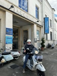 Person mit Helm sitzt auf einem Roller vor einem Gebäude mit Schildern, weitere Roller sind daneben geparkt.