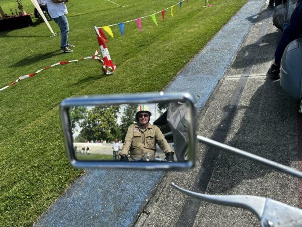 Motorradfahrer mit Helm und Jacke spiegelt sich im Rückspiegel, im Hintergrund Wiese mit Absperrband und eine Person.