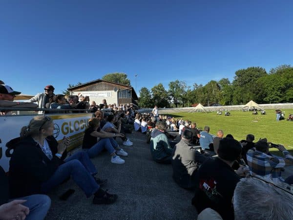 Viele Menschen sitzen auf einer Tribüne im Freien und schauen auf ein Spielfeld bei sonnigem Wetter.