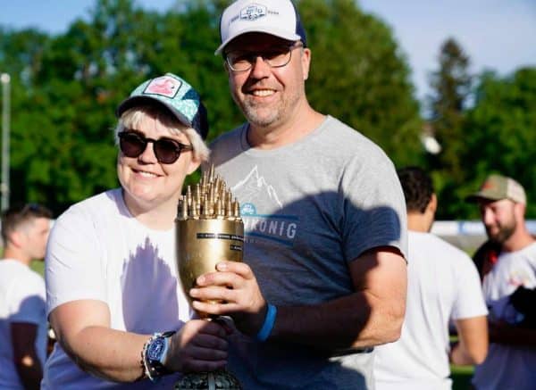 Zwei Personen stehen im Freien und halten gemeinsam einen goldenen Pokal, beide lächeln und tragen Sonnenbrillen und Kappen.