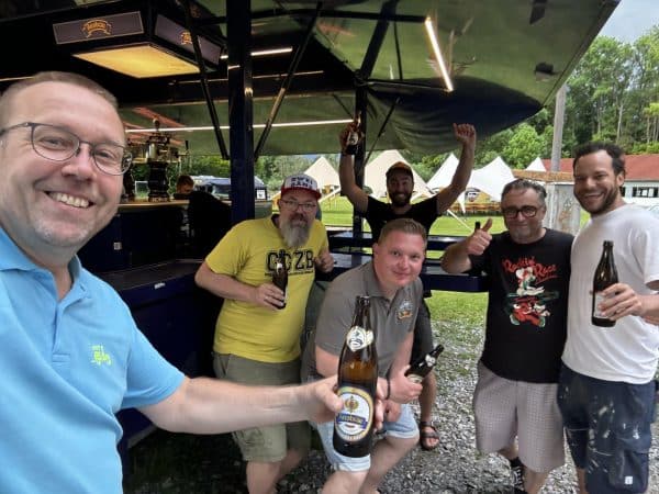 Fünf Männer stehen draußen zusammen, halten Bierflaschen in der Hand und lächeln in die Kamera, im Hintergrund Zelte und ein Imbisswagen.