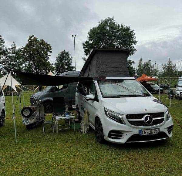 Weißer Mercedes-Camper mit geöffnetem Aufstelldach und Markise, Campingausstattung und Tisch im Freien, parkend auf einer Wiese bei bewölktem Himmel.