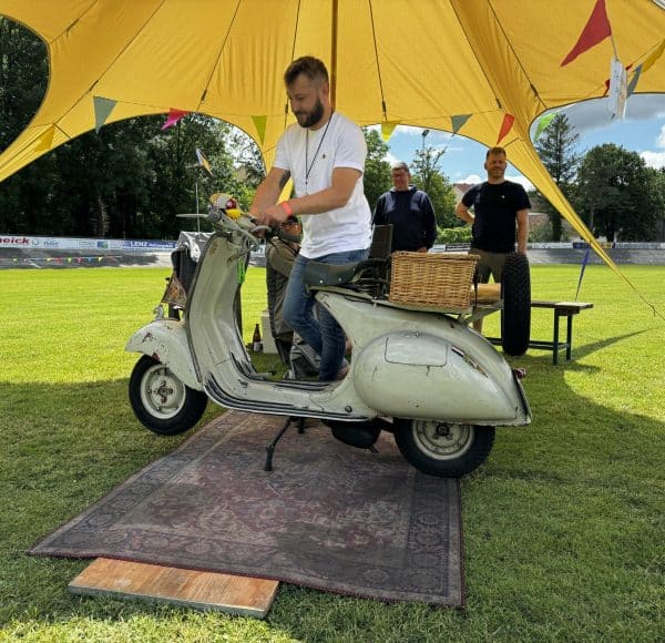 Mann steht auf einem weißen Oldtimer-Roller unter einem gelben Zelt auf einer Wiese, zwei weitere Personen im Hintergrund.