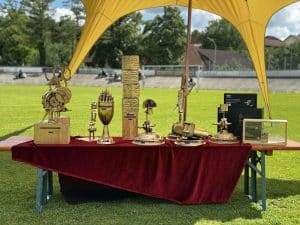 Ein Tisch mit roten Tuch, darauf mehrere kunstvolle Pokale und Trophäen im Freien auf einem Sportplatz ausgestellt.