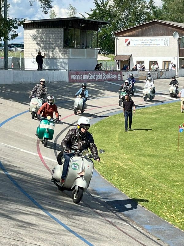 Mehrere Personen fahren auf klassischen Motorrollern auf einer Radrennbahn. Einige Zuschauer stehen am Rand und beobachten das Rennen bei sonnigem Wetter.