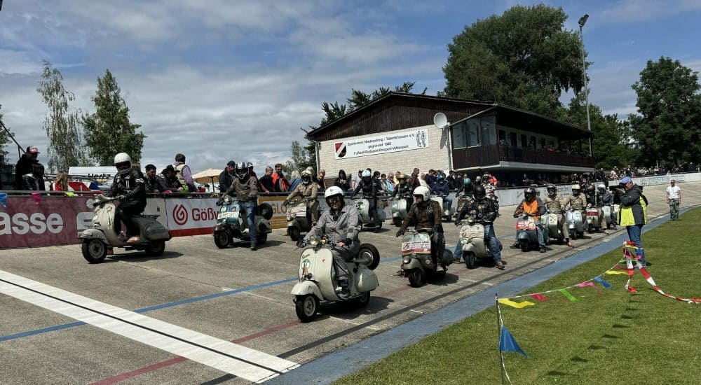 Mehrere Menschen fahren klassische Motorroller auf einer Rennstrecke bei einer Veranstaltung, mit Zuschauern und einem Gebäude im Hintergrund.