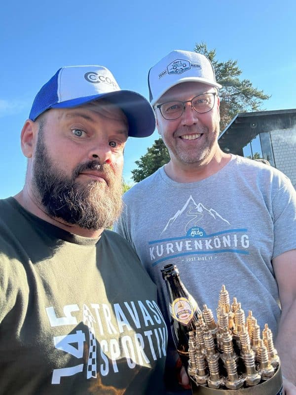 Zwei Männer mit Bärten und Caps stehen draußen, einer hält eine Flasche Bier, sonniger Tag, beide lächeln in die Kamera.