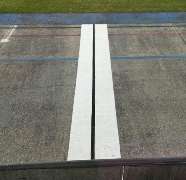 Zwei breite, weiße Linien auf Asphalt, vermutlich Teil einer Sportbahn, mit grünem Rasen im Hintergrund.