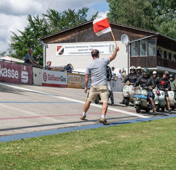 Ein Mann schwenkt eine rot-weiße Flagge, während mehrere Personen mit Motorrollern an einer Startlinie vor einem Gebäude stehen.
