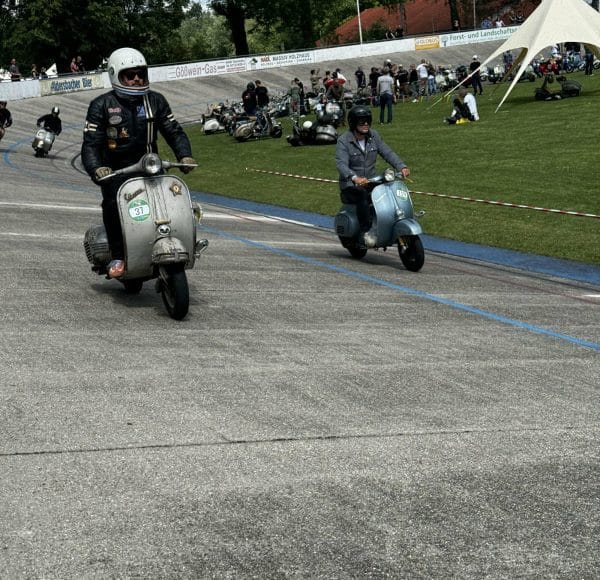 Mehrere Personen fahren alte Motorroller auf einer Rennstrecke im Freien, während Zuschauer auf dem Gras im Hintergrund stehen und sitzen.