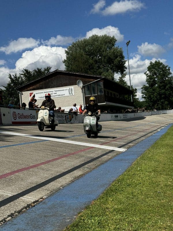 Zwei Personen fahren Motorroller auf einer Rennstrecke bei sonnigem Wetter, starten gerade ein Rennen. Im Hintergrund steht ein Gebäude und Bäume.
