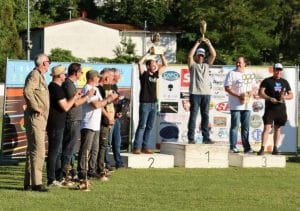 Mehrere Männer stehen auf einem Siegerpodest im Freien, heben Trophäen. Umstehende applaudieren, Banner mit Logos im Hintergrund.