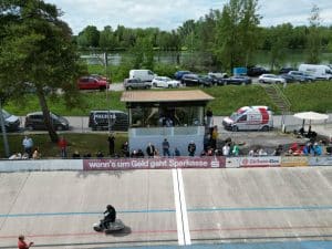 Radrennbahn mit Motorradfahrer auf der Bahn, Zuschauer und Fahrzeuge im Hintergrund, sonniger Tag.