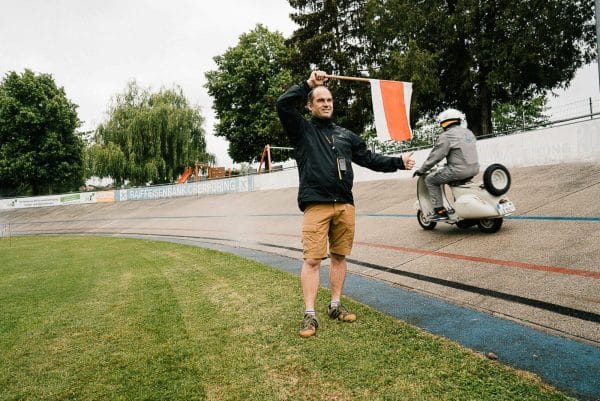 Ein Mann hält eine rot-weiße Flagge, während ein Rollerfahrer auf einer Rennbahn vorbeifährt; Bäume im Hintergrund.