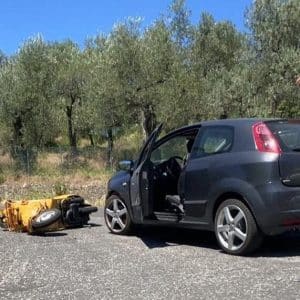 Schwarzes Auto mit offener Tür steht neben umgefallenem gelben Motorrad auf einer Landstraße, im Hintergrund Bäume und blauer Himmel.
