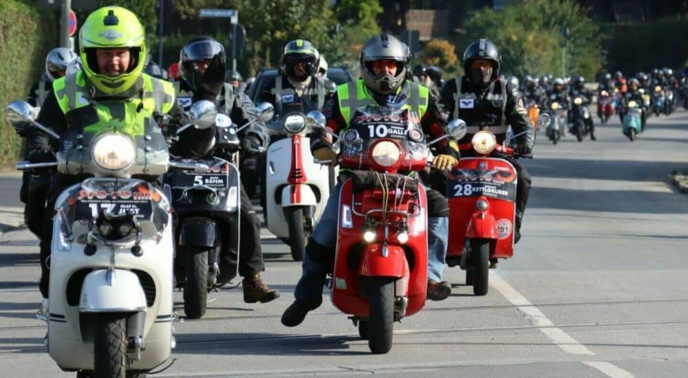 Gruppe von Menschen fährt auf Motorrollern mit Helmen auf einer Straße, vorne ein roter Roller, sonniges Wetter.
