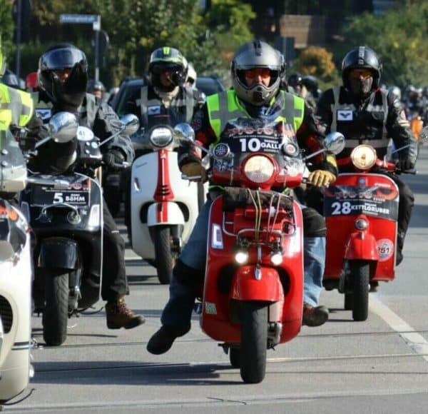 Gruppe von Menschen fährt auf Motorrollern mit Helmen auf einer Straße, vorne ein roter Roller, sonniges Wetter.