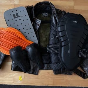 Motorradjacke auf Holzboden mit verschiedenen Protektoren und Schutzausrüstungen aus Kunststoff und Schaumstoff, in unterschiedlichen Größen und Formen, darüber verteilt.