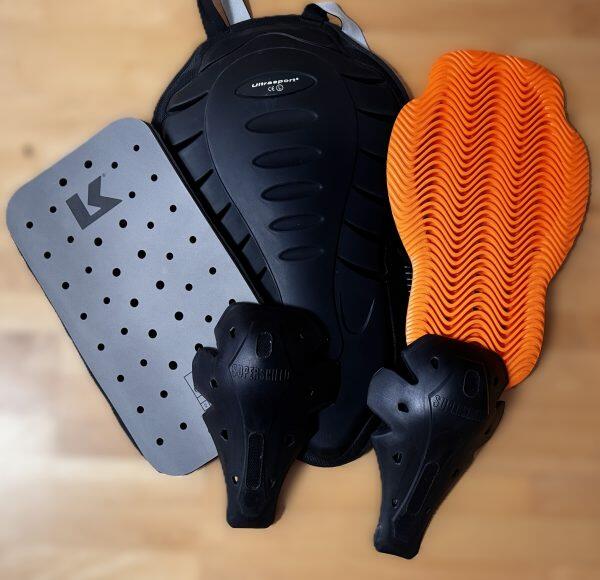 Drei verschiedene Rückenprotektoren für Motorradbekleidung, flach auf einem Holzboden ausgelegt, in Grau, Schwarz und Orange.