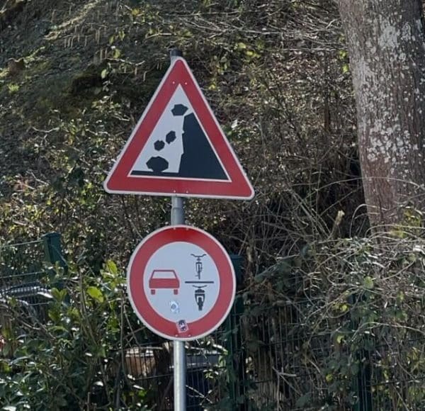 Zwei Verkehrsschilder: eines warnt vor Steinschlag, das andere verbietet Autos und Motorräder die Durchfahrt.