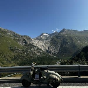 Roller steht vor einer malerischen Berglandschaft mit grünem Tal und schneebedeckten Gipfeln unter klarem, blauem Himmel.