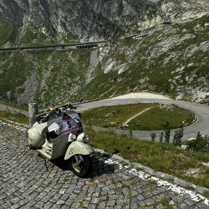Ein Motorroller steht auf Kopfsteinpflaster vor einer kurvigen Bergstraße, umgeben von grünen Hängen und Felsen.