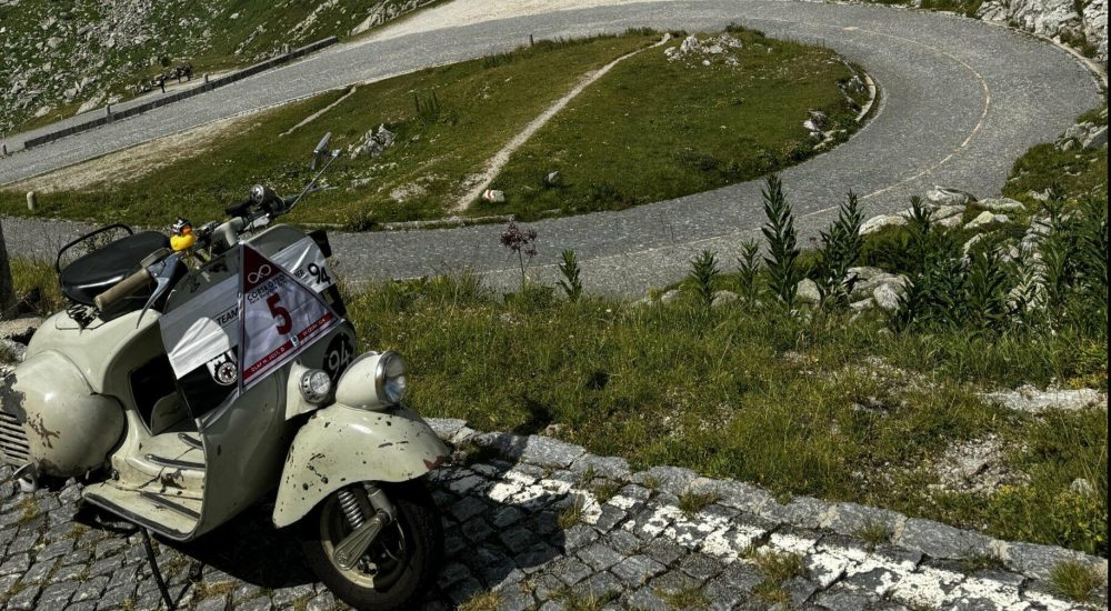 Ein weißer Motorroller steht auf Kopfsteinpflaster vor einer scharfen Kurve in den Bergen bei sonnigem Wetter.