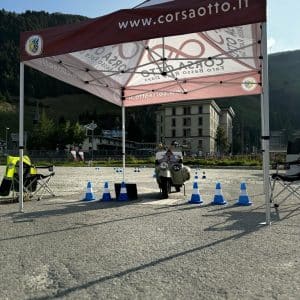 Motorradfahrer fährt unter einem Zelt mit „corsaosto.it“-Banner, umgeben von Verkehrshütchen auf einem Schotterplatz, Berge und Gebäude im Hintergrund.