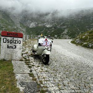 Oldtimer-Roller parkt auf einer kurvigen Kopfsteinpflasterstraße in den Bergen, daneben ein Straßenschild mit der Aufschrift „Ospizio km3“, Nebel im Hintergrund.