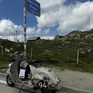 Alte Vespa steht an einem Straßenschild in den Bergen, umgeben von grüner Landschaft und bewölktem Himmel.